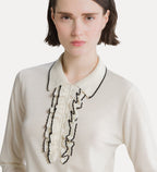 A close-up of a model wearing a cream-colored knit top with a ruffled collar and front placket outlined in black contrast stitching.