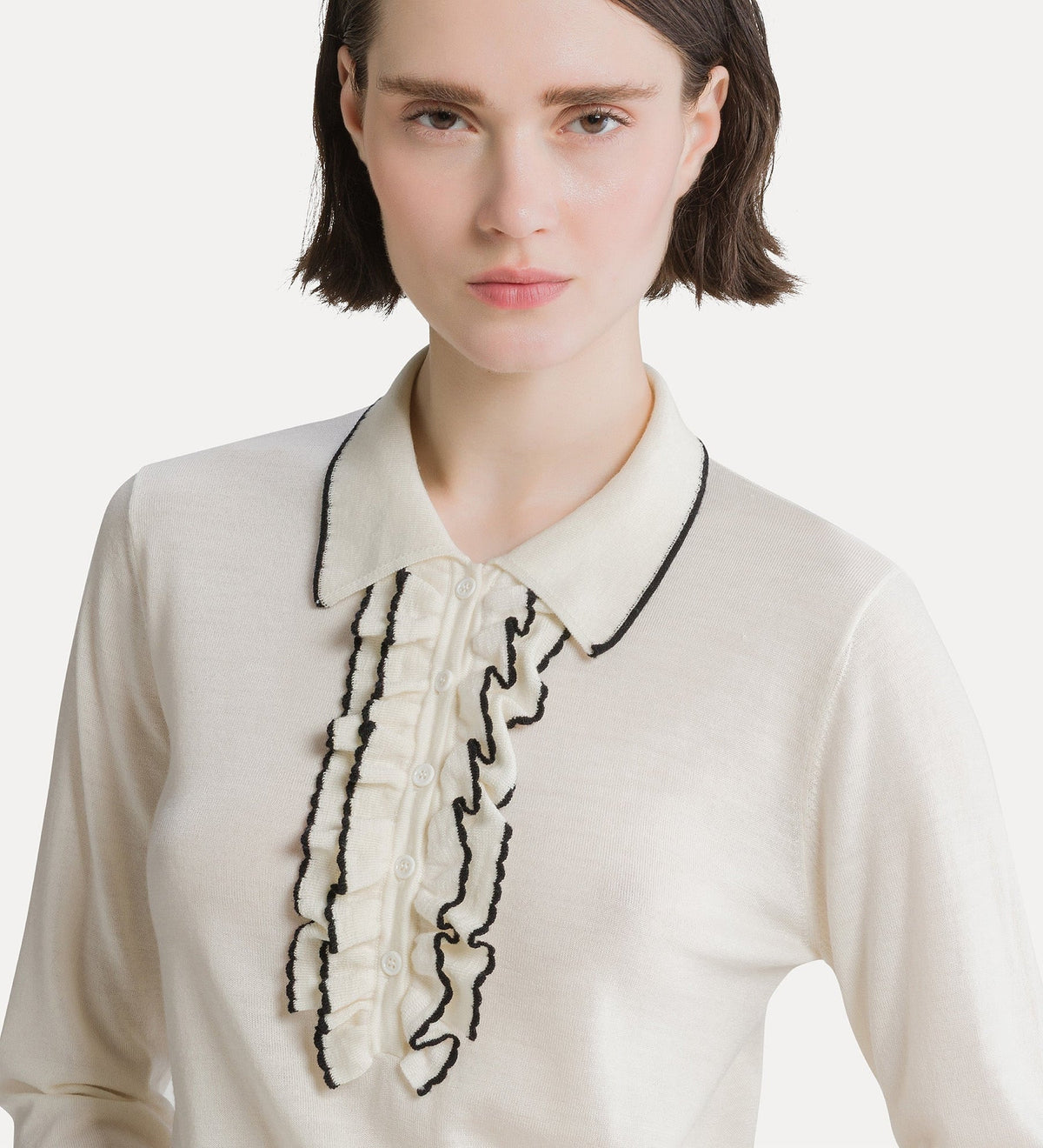 A close-up of a model wearing a cream-colored knit top with a ruffled collar and front placket outlined in black contrast stitching.