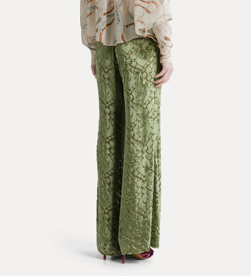 A person shown from the back wearing wide-leg sage green lace pants with a floral vine pattern, paired with a patterned blouse and burgundy heels, against a light gray studio background.