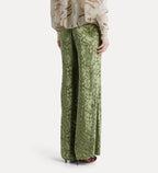 A person shown from the back wearing wide-leg sage green lace pants with a floral vine pattern, paired with a patterned blouse and burgundy heels, against a light gray studio background.