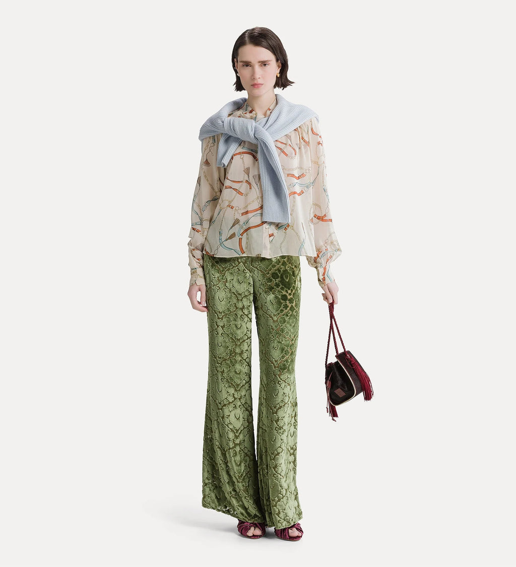 A model stands against a light gray studio background wearing a beige patterned blouse with a light blue draped scarf, green snakeskin-print wide-leg pants, and holding a small burgundy handbag.