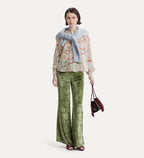 A model stands against a light gray studio background wearing a beige patterned blouse with a light blue draped scarf, green snakeskin-print wide-leg pants, and holding a small burgundy handbag.