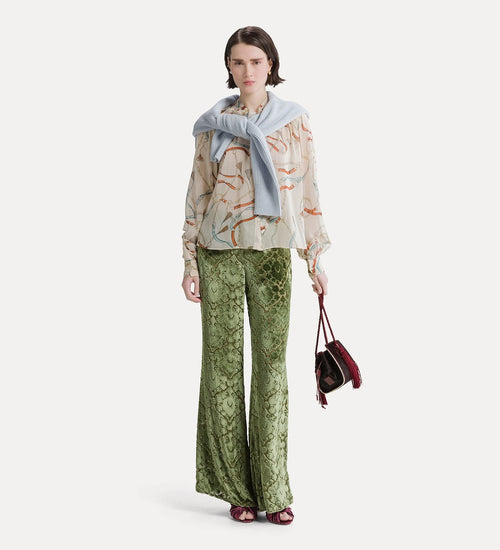 A model stands against a light gray studio background wearing a beige patterned blouse with a light blue draped scarf, green snakeskin-print wide-leg pants, and holding a small burgundy handbag.