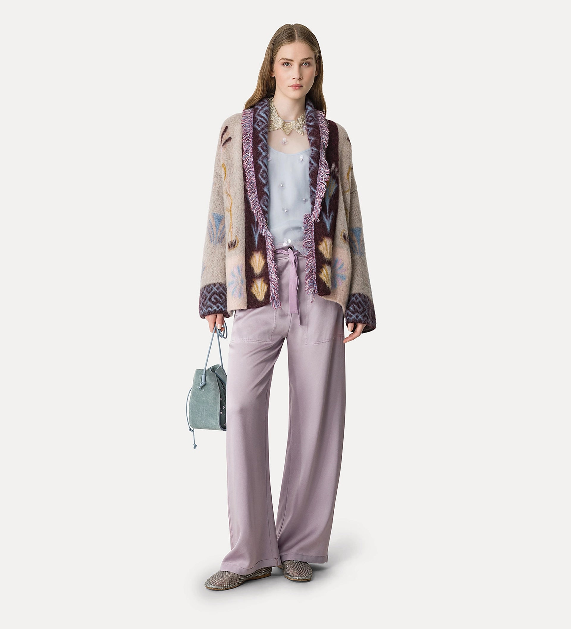 Woman wearing a patterned jacket, light purple pants, and holding a handbag on a white background