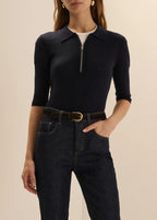 A woman wearing a dark navy ribbed knit polo-neck top with a short front zipper, paired with dark blue jeans and a brown belt, against a plain beige studio background.