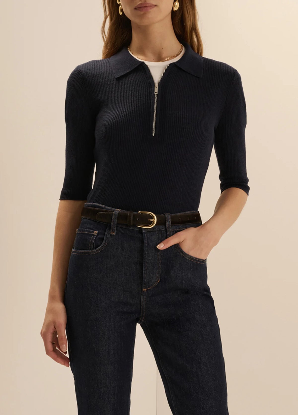 A woman wearing a dark navy ribbed knit polo-neck top with a short front zipper, paired with dark blue jeans and a brown belt, against a plain beige studio background.