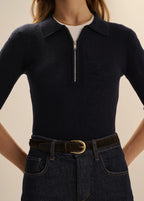 A close-up of a model wearing a dark navy ribbed knit polo-neck top with a short front zipper, tucked into dark blue jeans with a brown belt.
