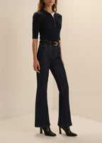 A model wearing a dark navy blue half-zip mock-neck top, dark blue high-waisted flare jeans with a black belt, and black heeled boots, standing against a warm beige studio background.