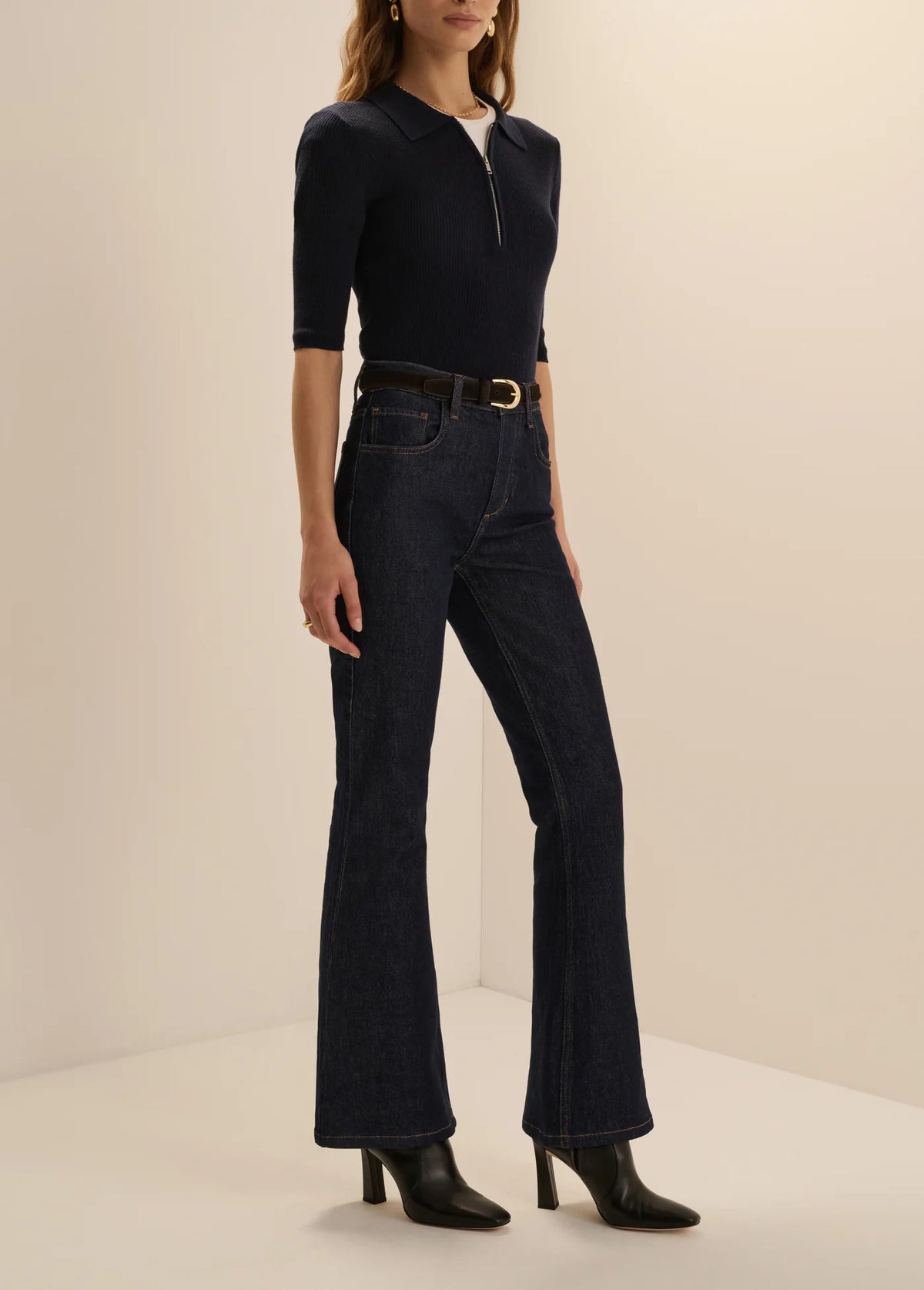 A model wearing a dark navy blue half-zip mock-neck top, dark blue high-waisted flare jeans with a black belt, and black heeled boots, standing against a warm beige studio background.