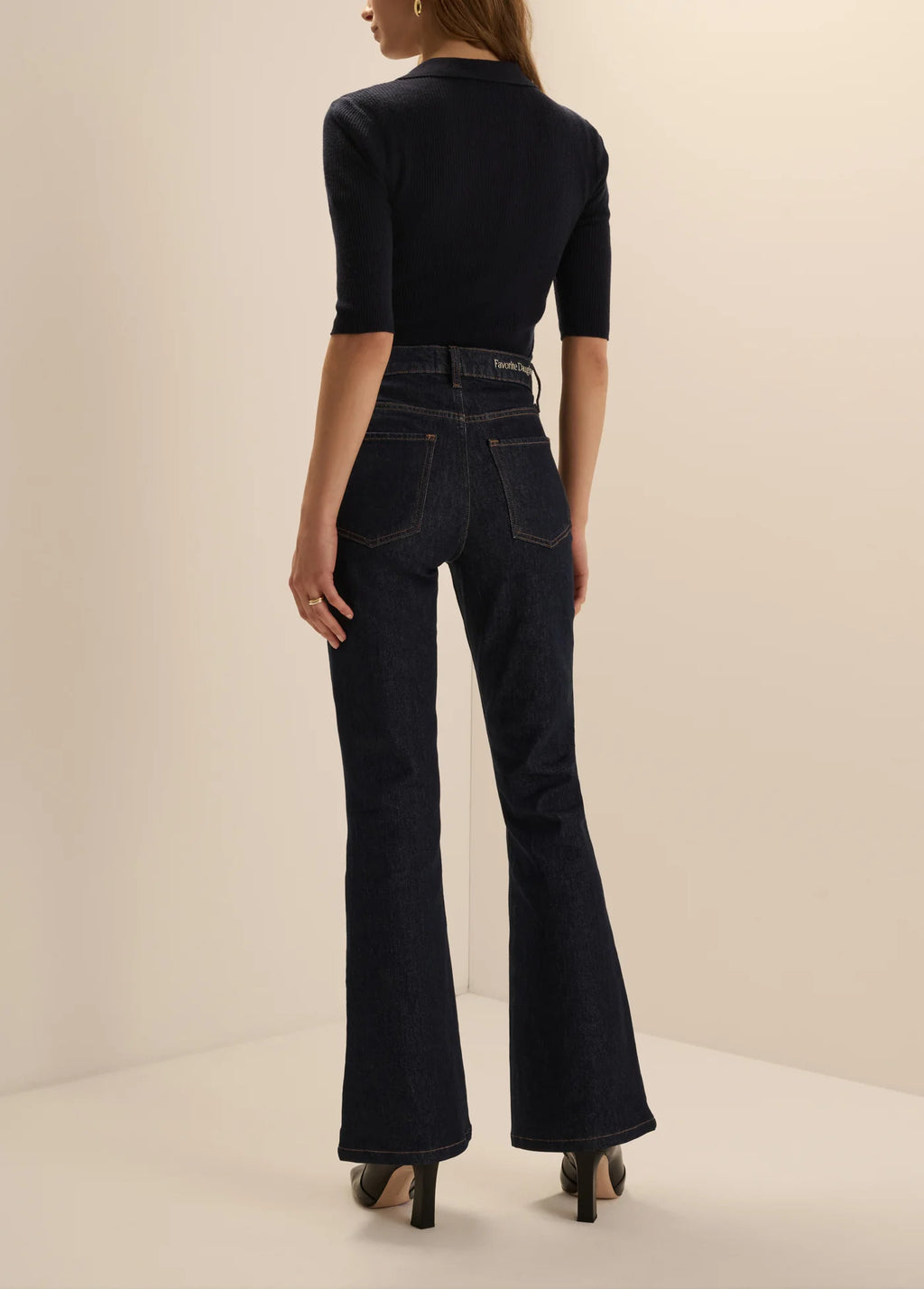 A model shown from the back wearing dark navy blue high-waisted flare jeans and a matching dark navy short-sleeve top, standing against a plain beige studio background.