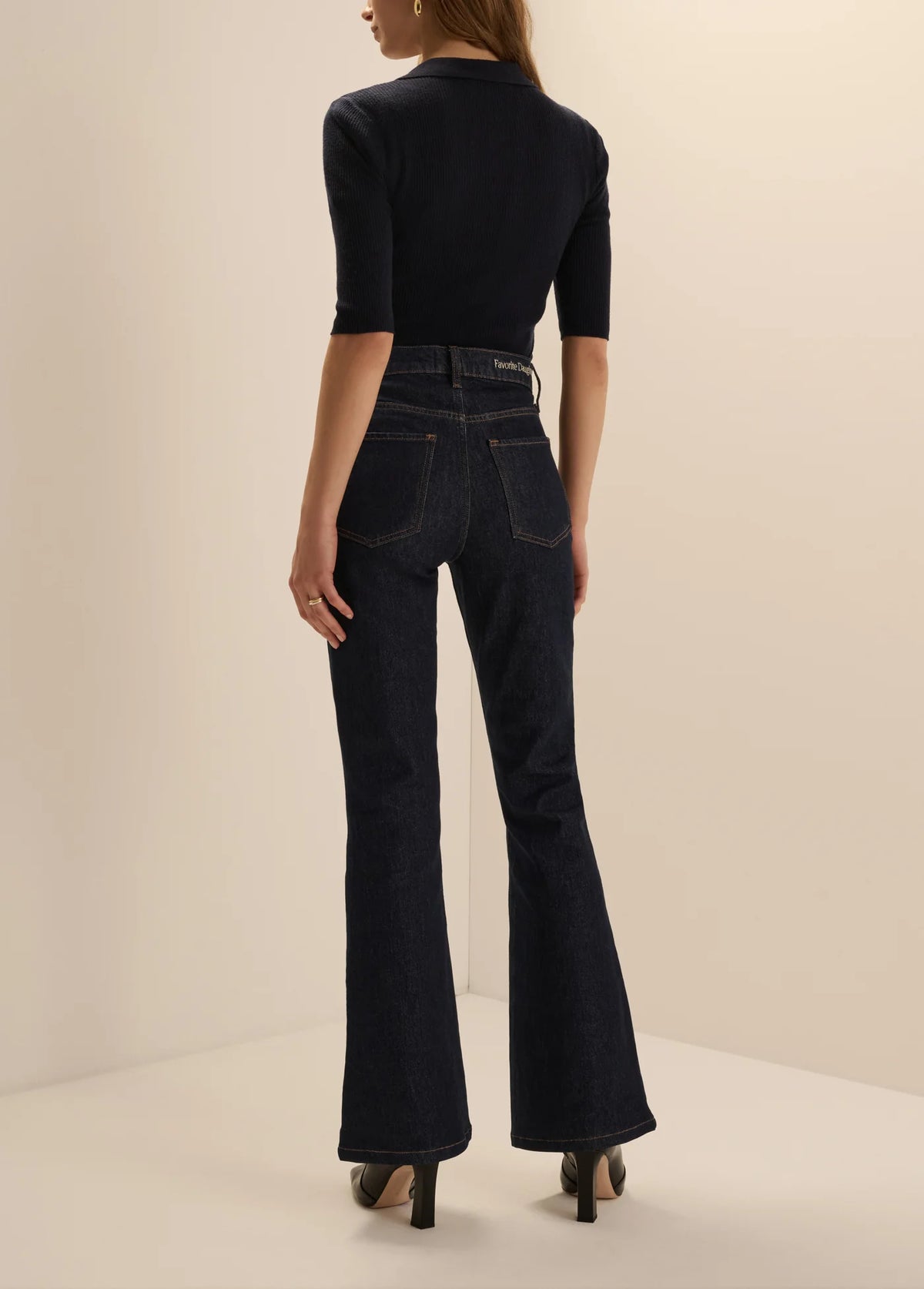 A model shown from the back wearing dark navy blue high-waisted flare jeans and a matching dark navy short-sleeve top, standing against a plain beige studio background.