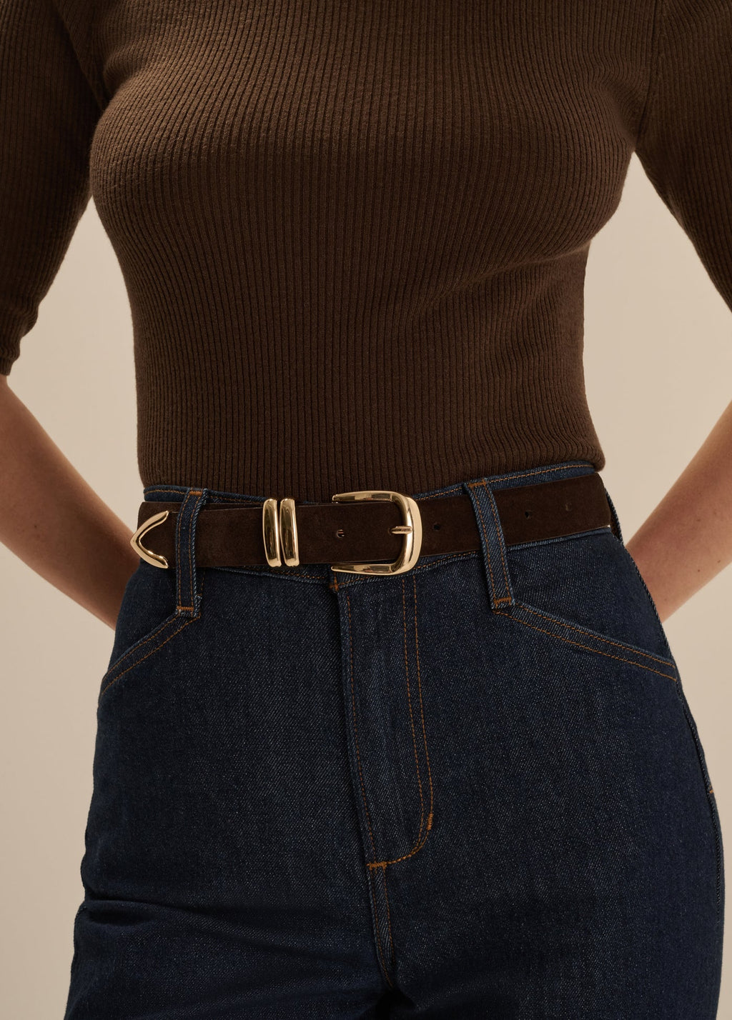 A close-up of a model wearing a dark brown ribbed knit top, dark blue jeans, and a dark brown leather belt with a gold-tone buckle.