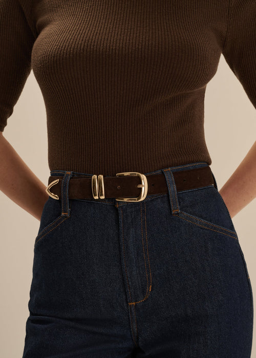 A close-up of a model wearing a dark brown ribbed knit top, dark blue jeans, and a dark brown leather belt with a gold-tone buckle.