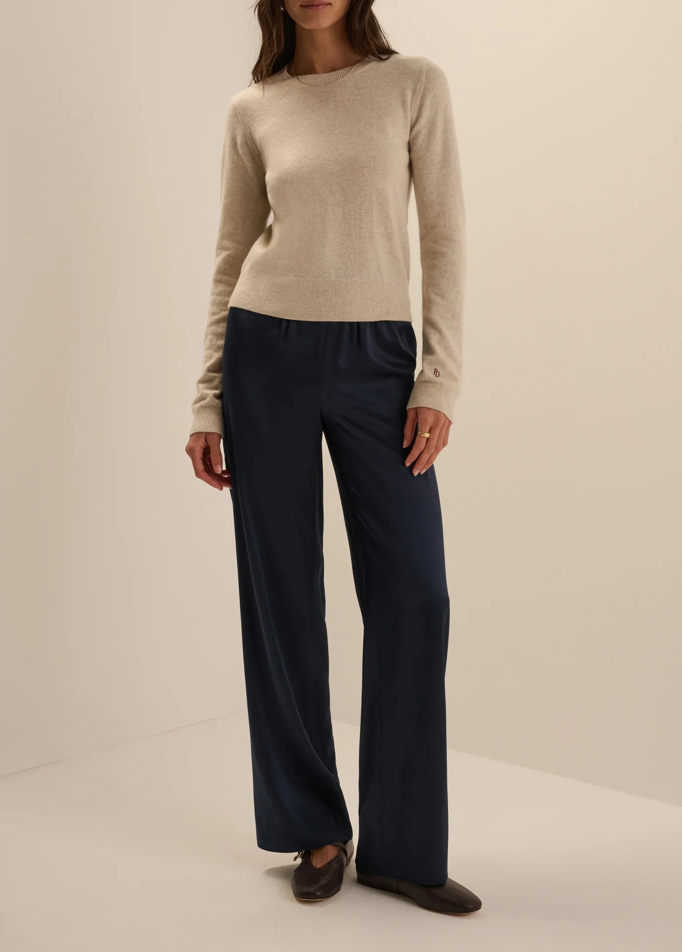 A model wearing a beige knit sweater and dark navy wide-leg trousers, standing against a plain beige studio background.