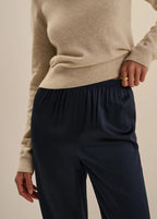 A close-up of a person wearing a beige knit sweater and dark navy satin trousers, with one hand pulling the elastic waistband of the pants.