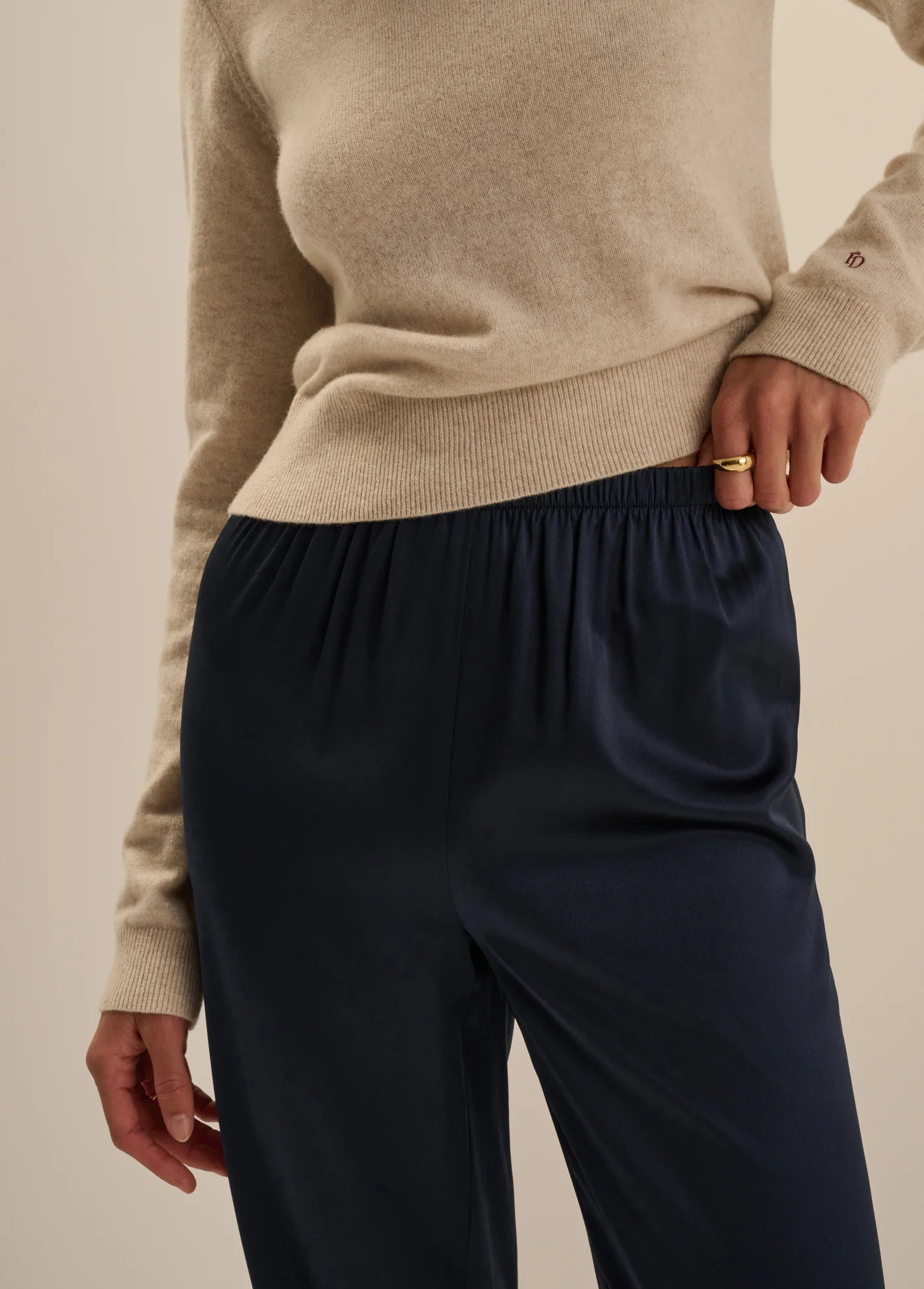 A close-up of a person wearing a beige knit sweater and dark navy satin trousers, with one hand pulling the elastic waistband of the pants.