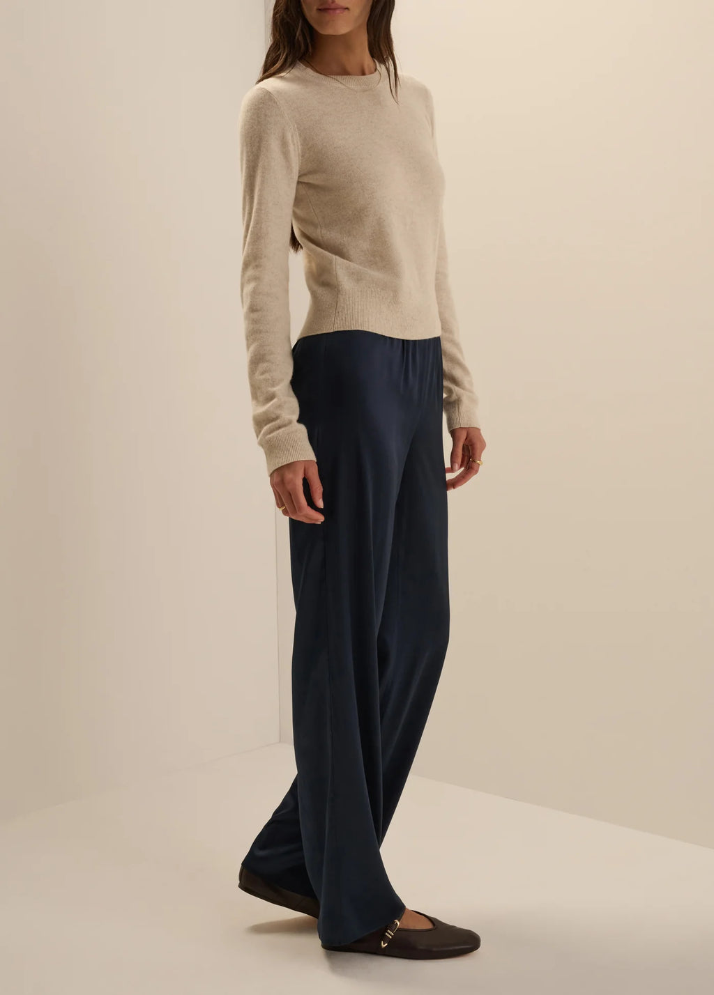 A woman modeling a cream knit long-sleeve top and dark navy wide-leg trousers, paired with dark brown flat shoes, in a studio setting with a warm off-white background.