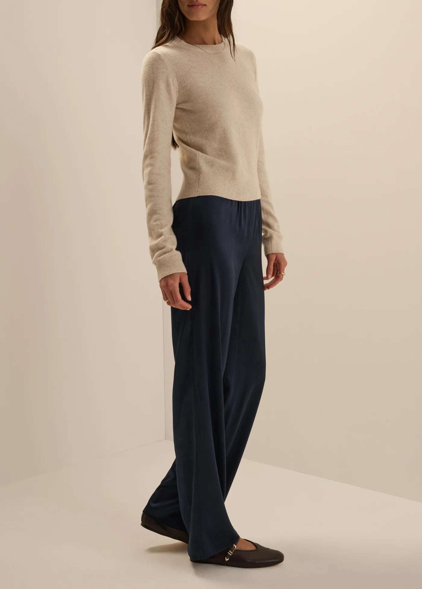 A woman modeling a cream knit long-sleeve top and dark navy wide-leg trousers, paired with dark brown flat shoes, in a studio setting with a warm off-white background.