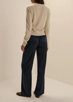 A woman shown from the back wearing a light beige knit sweater and dark navy wide-leg trousers, standing against a plain warm off-white studio background.