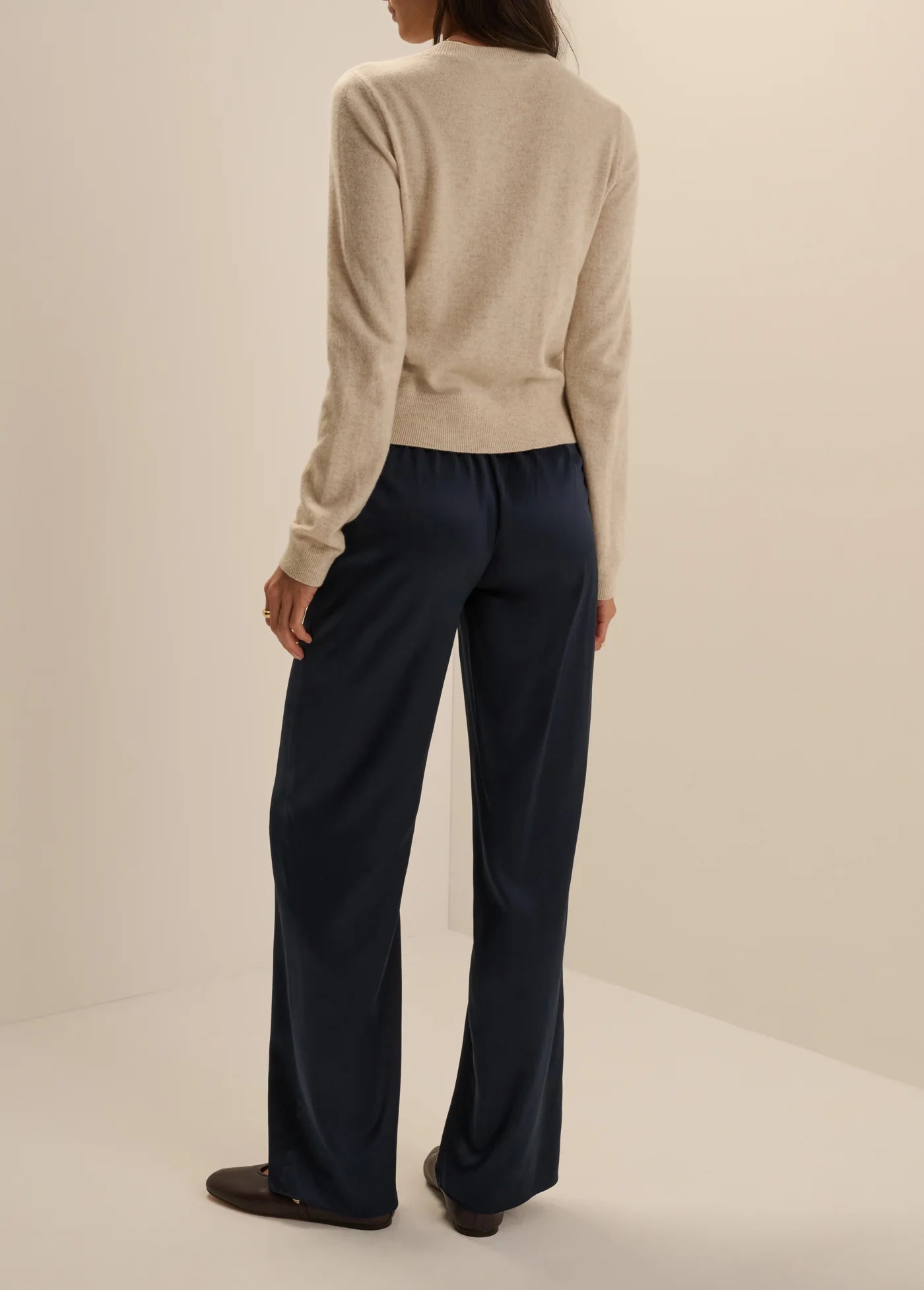 A woman shown from the back wearing a light beige knit sweater and dark navy wide-leg trousers, standing against a plain warm off-white studio background.
