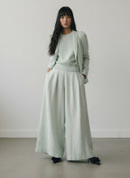 A woman stands in a minimalist studio wearing a light sage green outfit consisting of a long-sleeve top, an open blazer, and wide-leg pleated trousers, paired with black pointed shoes.

Hannes Roether