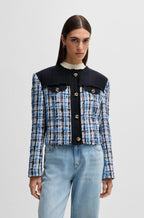 A female model wearing a tweed jacket with contrast panels and knot buttons in a check pattern.