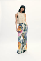 A model stands against a seamless white studio background wearing a sleeveless beige top with black buttons and wide-leg floral pants featuring orange, yellow, blue, and purple watercolor-style floral patterns.
