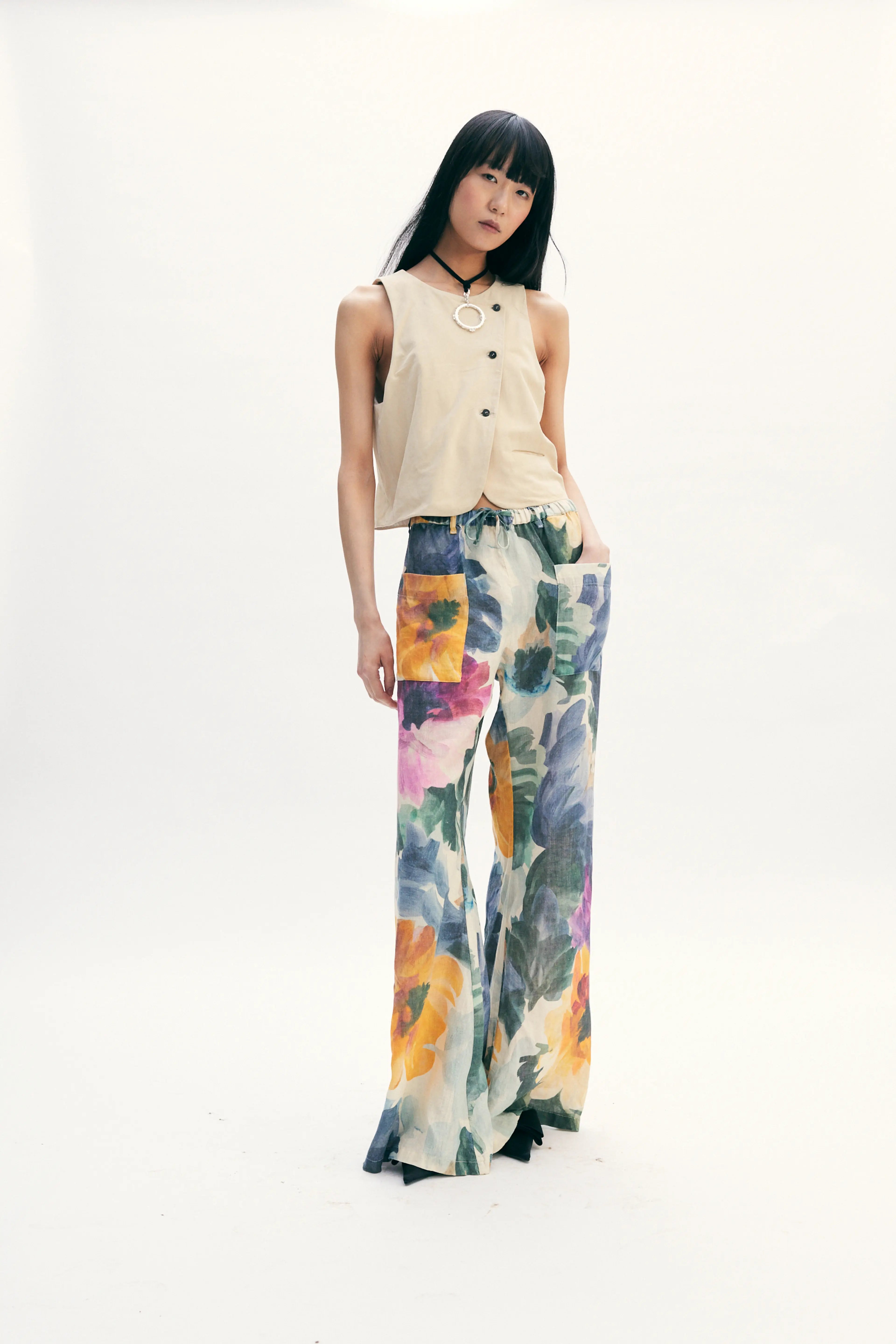 A model stands against a seamless white studio background wearing a sleeveless beige top with black buttons and wide-leg floral pants featuring orange, yellow, blue, and purple watercolor-style floral patterns.