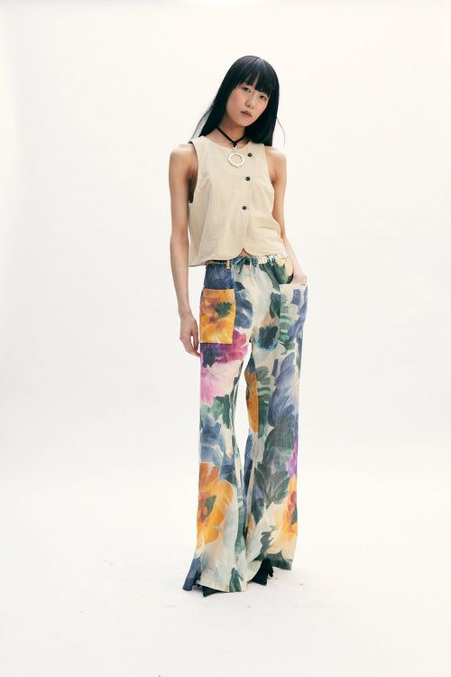 A model stands against a seamless white studio background wearing a sleeveless beige top with black buttons and wide-leg floral pants featuring orange, yellow, blue, and purple watercolor-style floral patterns.