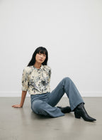 A woman sits on a concrete floor against a white wall, wearing a cream-colored blouse with a black and gray floral print, wide-leg blue jeans, and black ankle boots.
do29di.4059
Hannes Roether