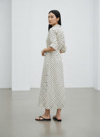 A woman stands in a minimalist white studio wearing a long-sleeved midi dress with a black polka dot pattern on a white background, paired with black flip-flop sandals.
bet28ter-k.4061
Hannes Roether
