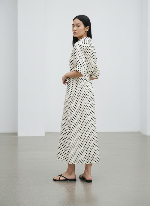A woman stands in a minimalist white studio wearing a long-sleeved midi dress with a black polka dot pattern on a white background, paired with black flip-flop sandals.
bet28ter-k.4061
Hannes Roether