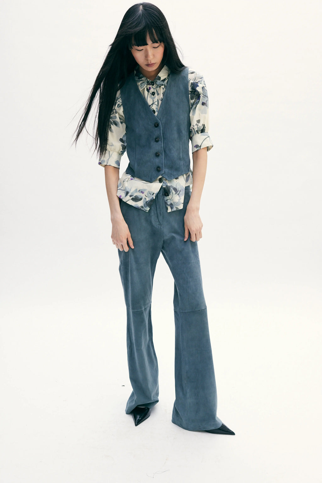 A model with long black hair is wearing a matching slate blue two-piece set consisting of a button-front vest and wide-leg pants, layered over a cream and blue floral print shirt, against a plain white studio background.
do29di.4059
Hannes Roether