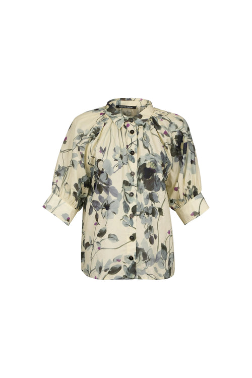 A women's short-sleeve blouse with a gathered, ruffled neckline and a watercolor-style floral print in muted gray, black, and soft pink on a cream background, shown against a light blue-gray studio background.

do29di.4059
hannes roether