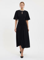 A woman modeling a black midi dress with short flutter sleeves, a keyhole neckline with a pearl-like collar, and a gathered ruched waist, photographed against a plain light gray studio background.