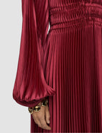 Red pleated dress with long sleeves and a hand wearing a gold bracelet on a light gray background
