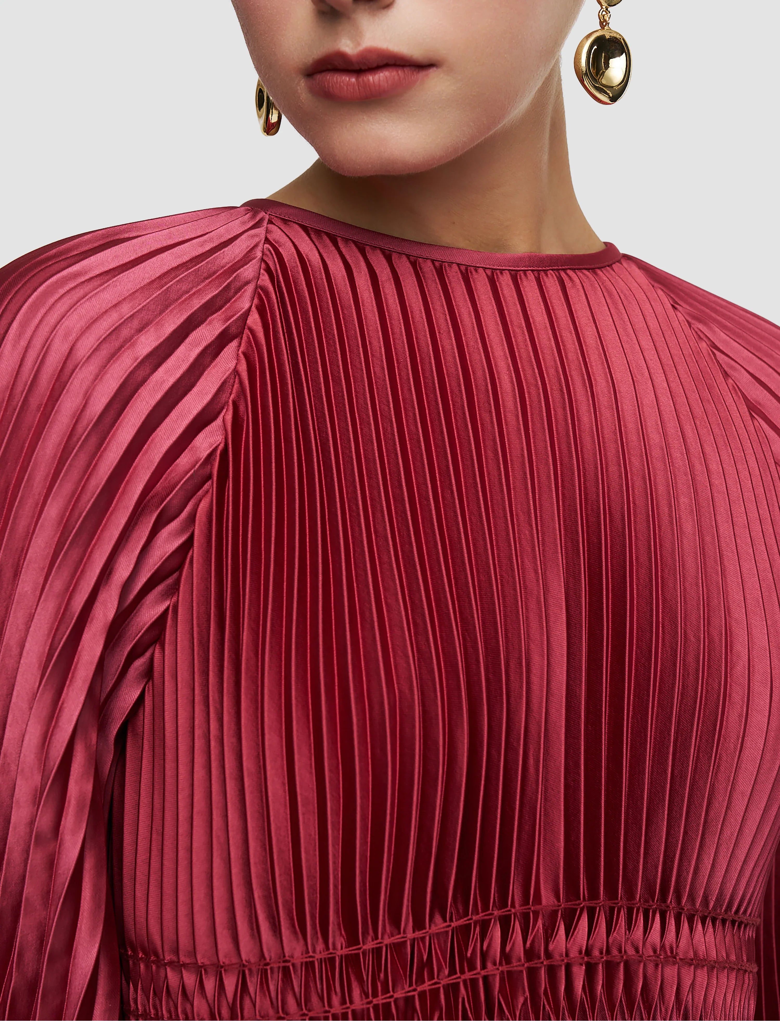 Close-up of a person wearing a pleated red dress with gold earrings on a light gray background