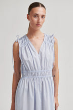 A woman wearing a sleeveless light blue and white vertically striped dress with a smocked waist and tie details on the shoulders, photographed against a neutral light gray studio background.