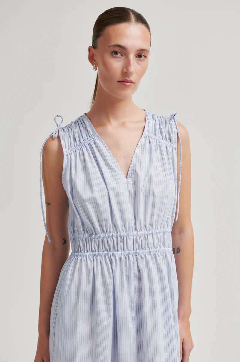 A woman wearing a sleeveless light blue and white vertically striped dress with a smocked waist and tie details on the shoulders, photographed against a neutral light gray studio background.