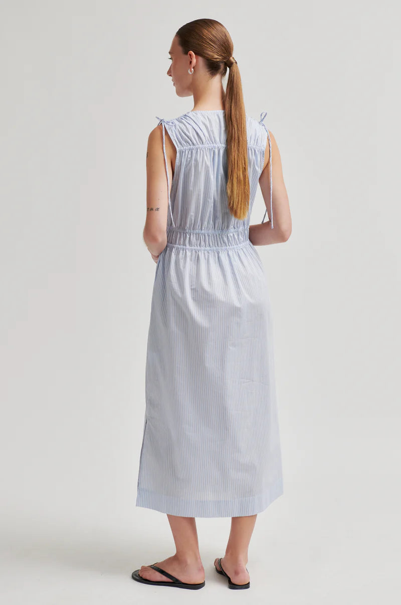 A woman shown from the back wearing a sleeveless light blue and white vertically striped midi dress with a smocked waist and tie straps at the shoulders, paired with black flip-flop sandals, against a plain light gray studio background.