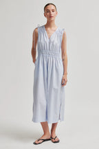 A woman modeling a light blue sleeveless midi dress with shoulder tie details and a smocked waist, paired with black flip-flop sandals, against a neutral light gray studio background.