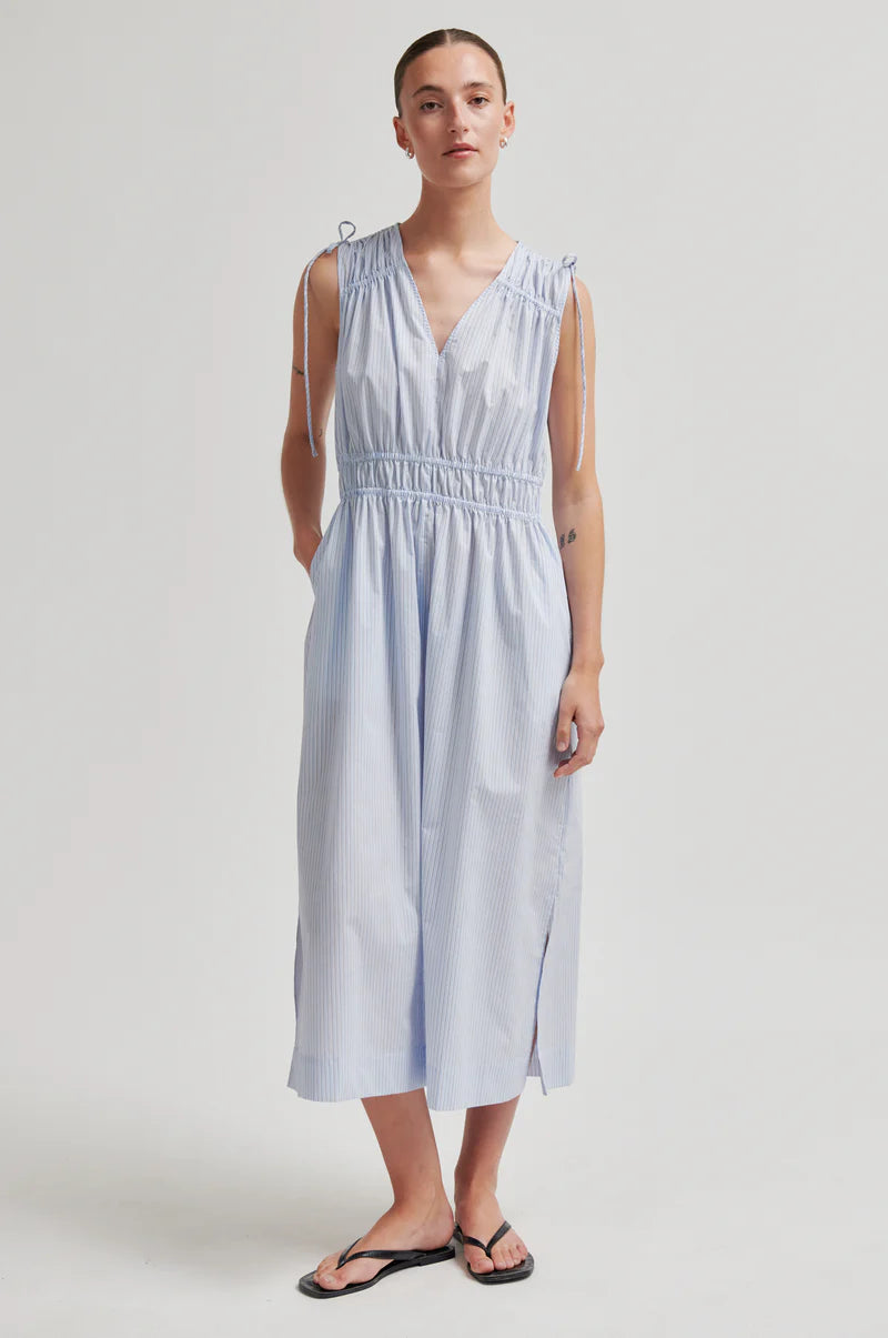 A woman modeling a light blue sleeveless midi dress with shoulder tie details and a smocked waist, paired with black flip-flop sandals, against a neutral light gray studio background.