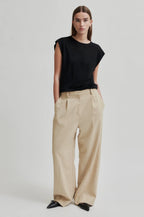 A female model wearing beige melange pleated trousers with a black top. Accessories include shoes and a belt.