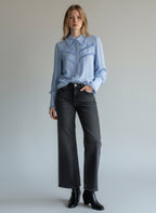 Woman wearing a light blue denim shirt and black jeans against a plain background