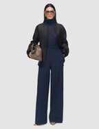 Woman wearing a black leather jacket, navy turtleneck, and wide-leg pants holding a beige handbag on a white background.